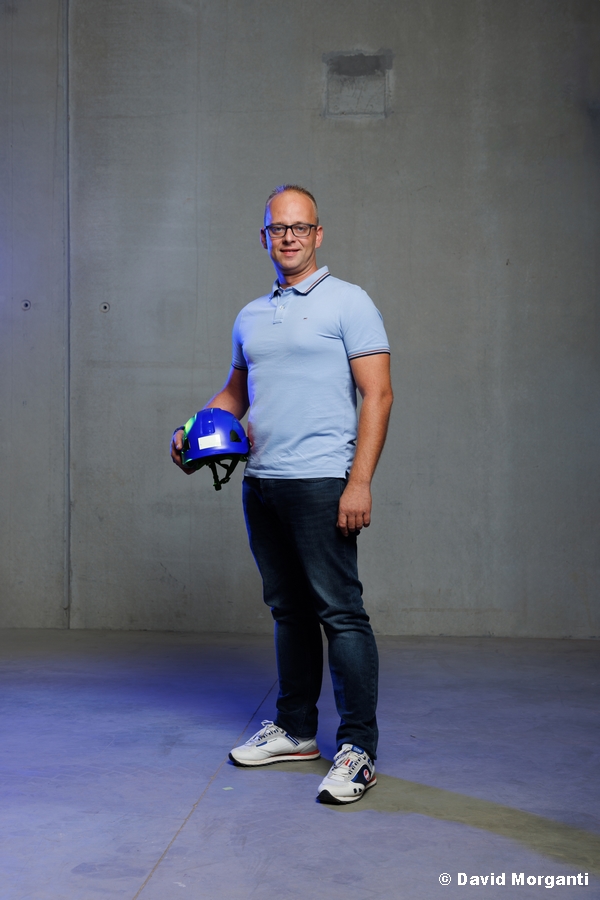 Portrait de Stéphane, directeur, souriant en polo bleu ciel et jean, tenant un casque de chantier bleu.